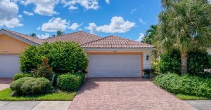 A tan house with two white garage doors and a stamped brick driveway bordered by green shrubs and palm trees.