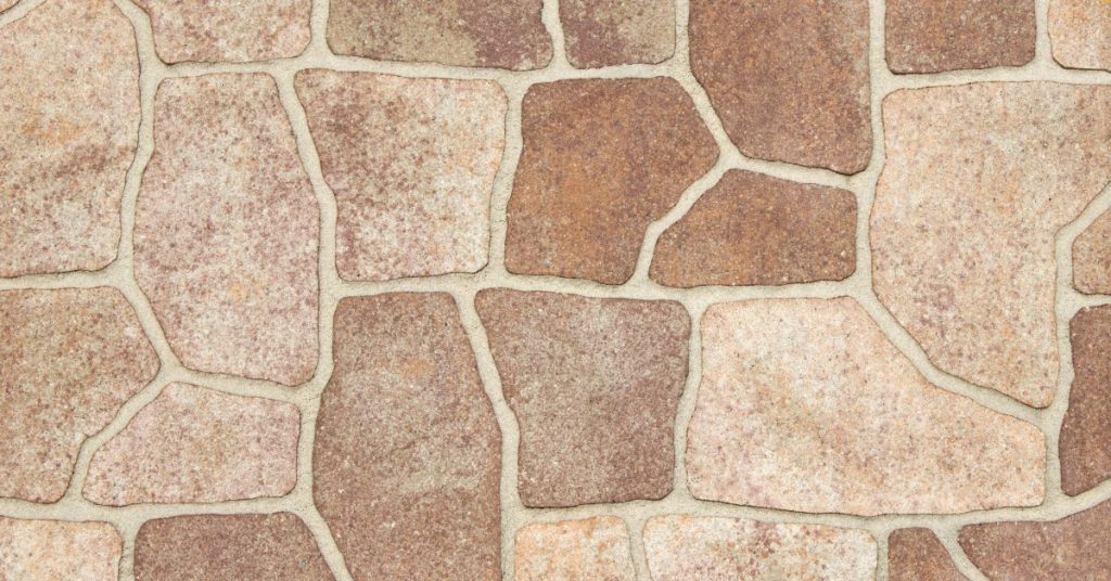 A pattern of irregular tan and brown stone with pale mortar joints and textured surfaces in varied shapes.