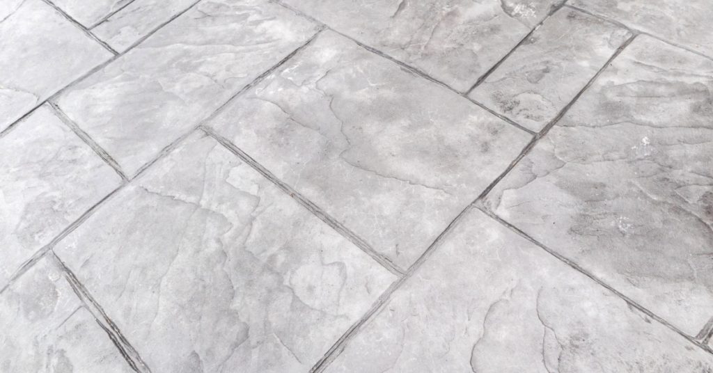 A gray stamped concrete surface with rectangular slab patterns, dark seams, mottled texture, and diagonal lines.
